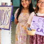 Two women warmly greet visitors at a community event in Ciudad de México with signs saying 'Bienvenidos' and 'Estás en Casa'.