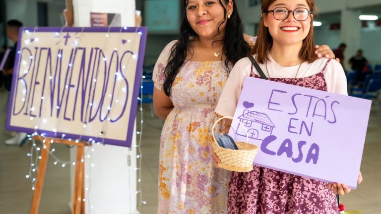 Two women warmly greet visitors at a community event in Ciudad de México with signs saying 'Bienvenidos' and 'Estás en Casa'.
